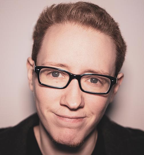 Autor Nick Böcker: Rote Haare unter einer schwarzen Beanie; ein breites Lächeln auf den Lippen; schwarzer Pulli